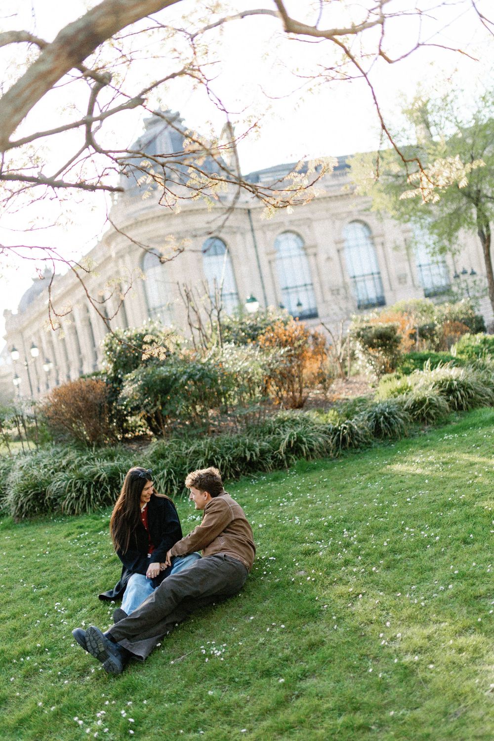 A couple relaxes on a grassy lawn in front of an ornate European-style building with arched windows.