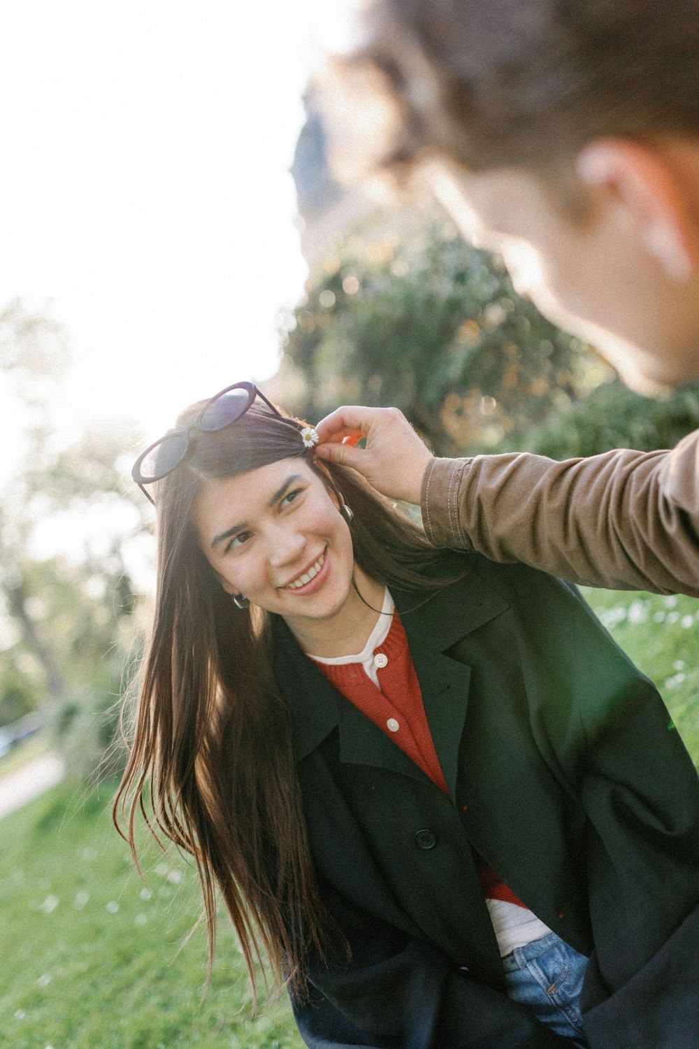 Someone wearing a green coat and red shirt smiles while having their sunglasses adjusted in a sunny outdoor setting.