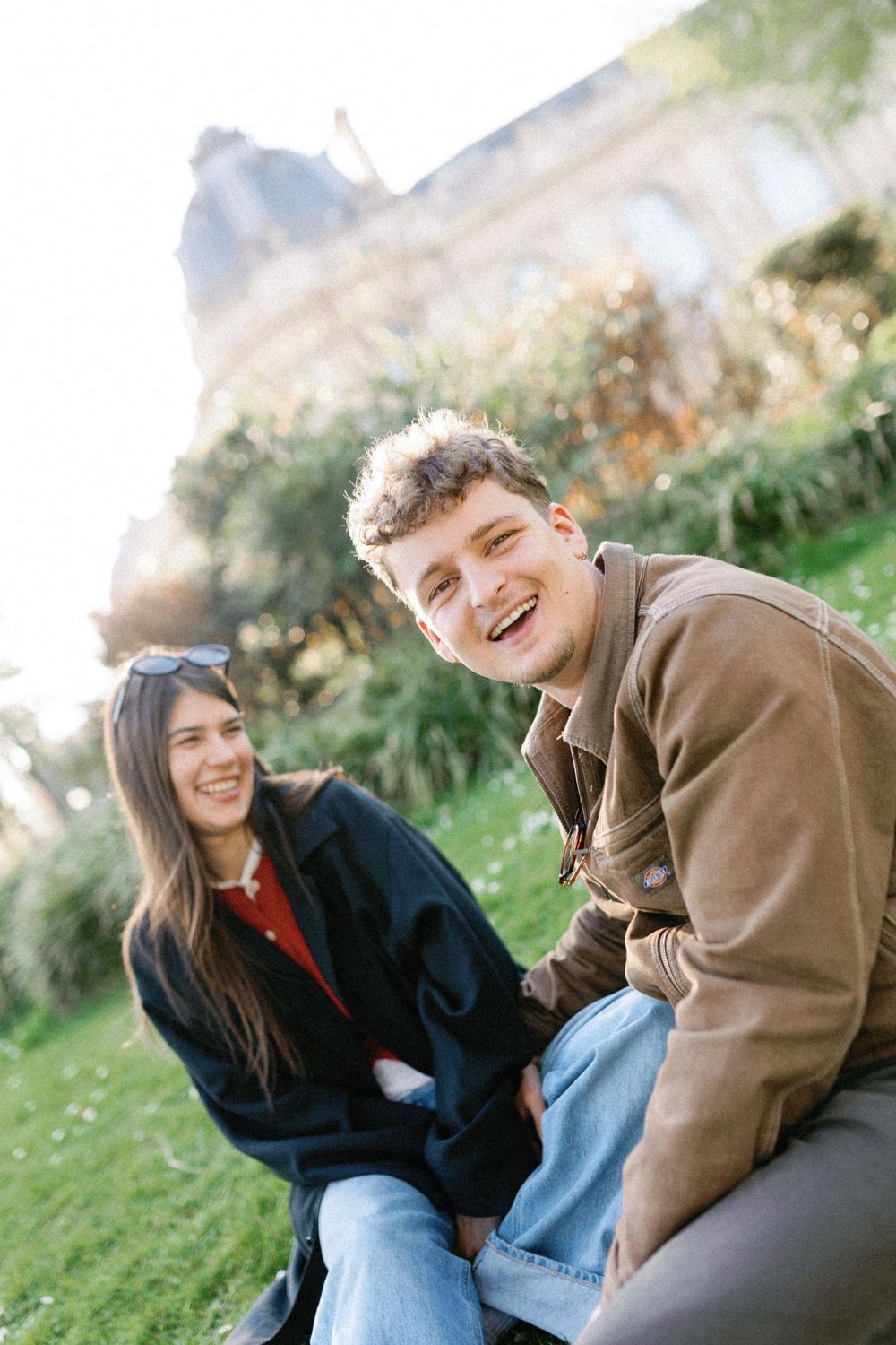 Two people share joyful laughter while sitting together on a grassy hillside on a sunny day.