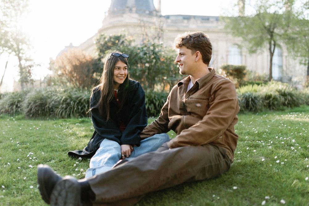 Two people sitting together on grass enjoying a relaxed conversation on a sunny day in a park setting.