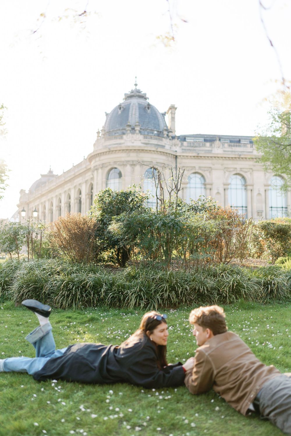 Couple laying on grass in front of elegant Parisian building with ornate architecture on sunny day.