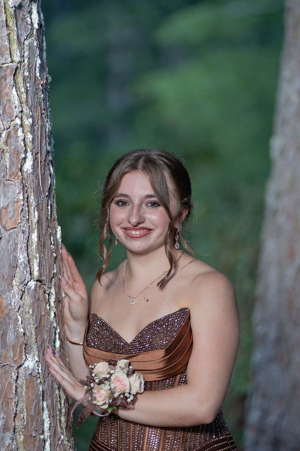 Young woman in sparkly bronze strapless prom dress with floral wrist corsage posing by tree in forest.