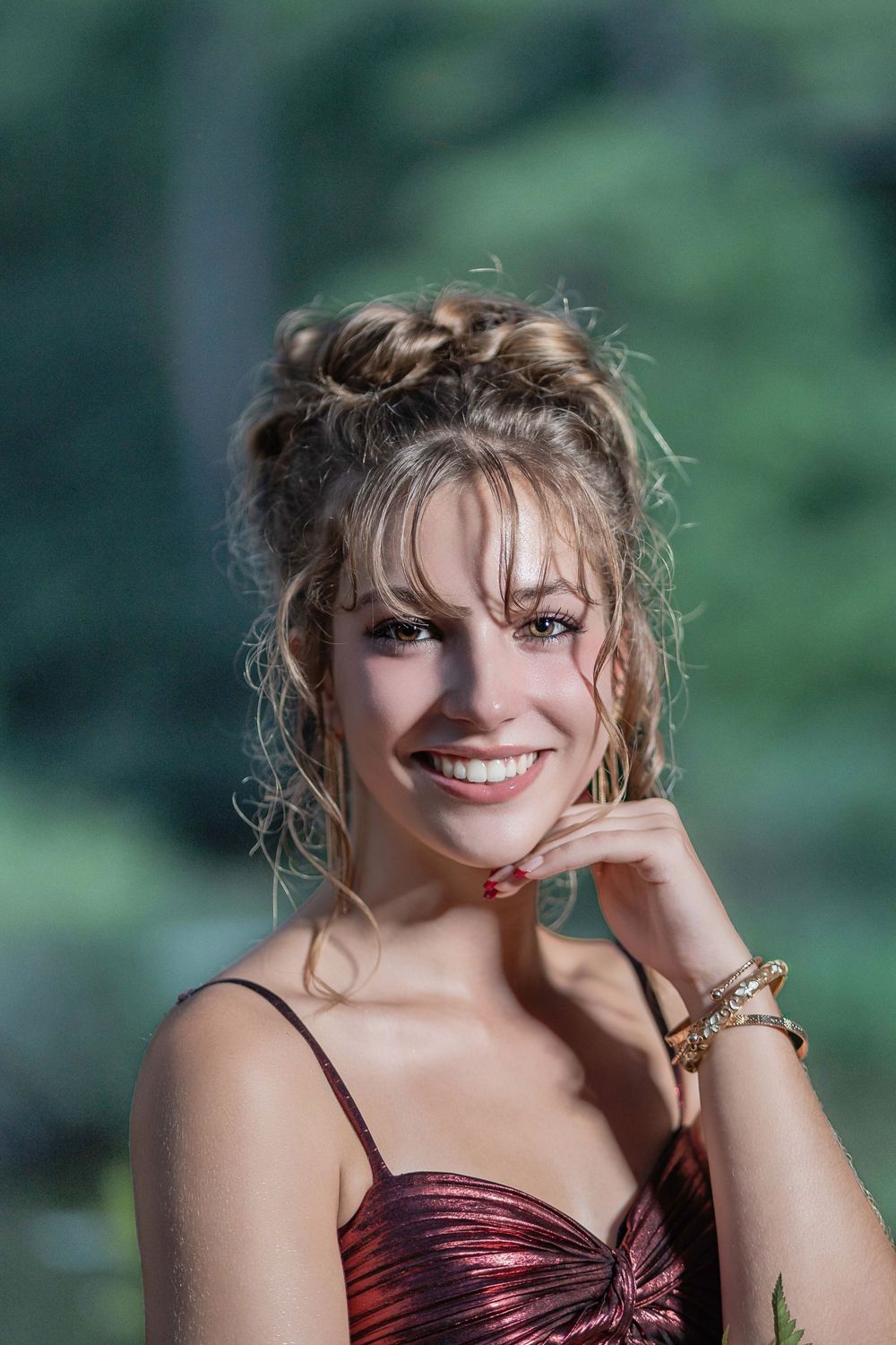 Smiling young woman in a red sparkly dress with an updo hairstyle and gold bracelet posing outdoors.
