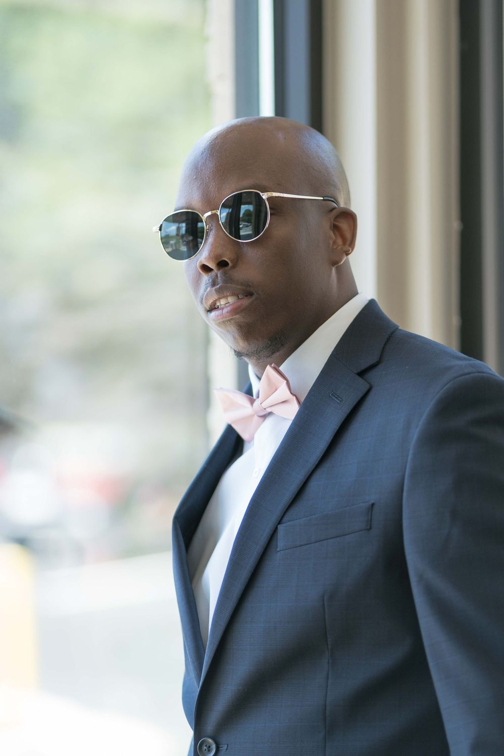 Stylish person in a dark suit and pink bow tie wearing sunglasses in natural window lighting.