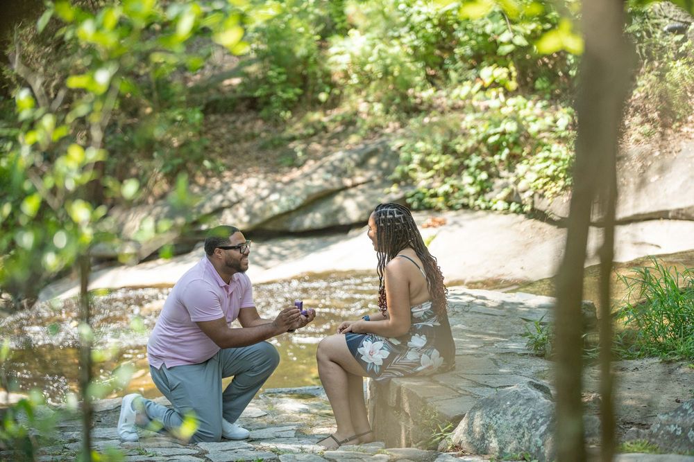 Man kneeling to propose to woman by a rocky creek in a lush green forest setting.