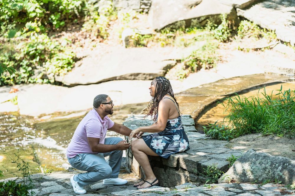 Man kneeling before woman on rocky riverbank in a romantic outdoor marriage proposal surrounded by lush greenery.