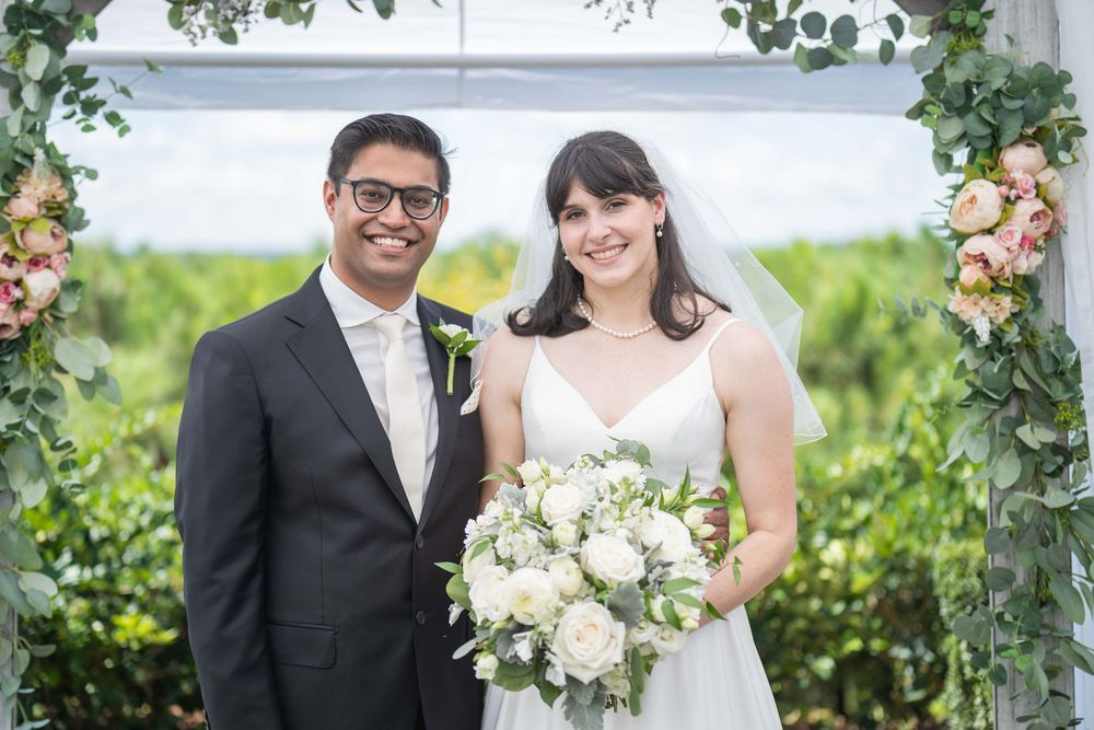 Wedding couple poses under floral arch in outdoor garden setting with white bouquet.
