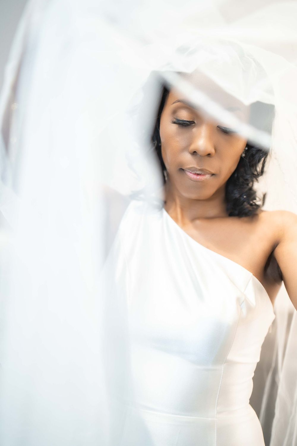 Ethereal bridal portrait through sheer veil featuring one-shoulder white wedding gown.
