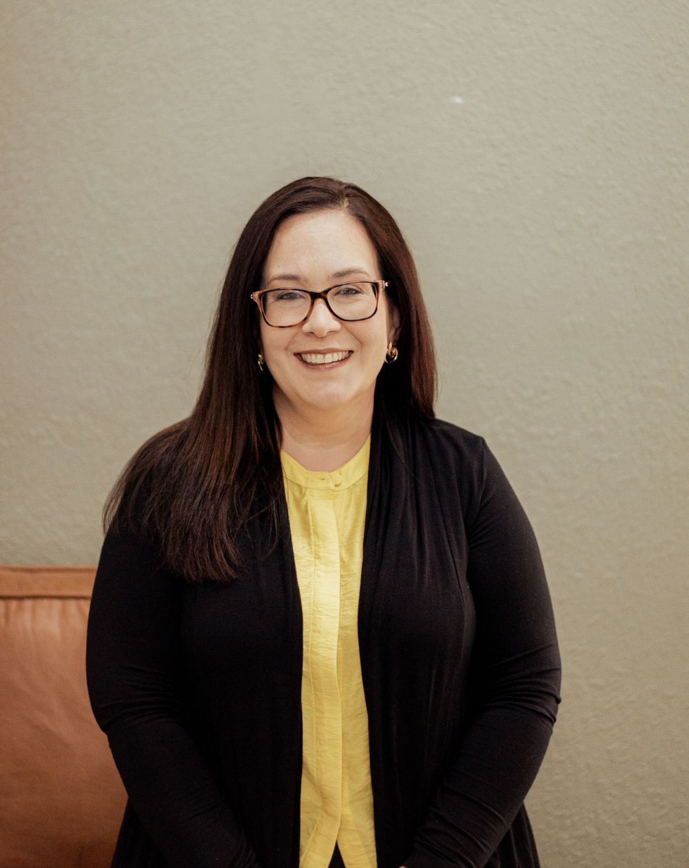Portrait against beige wall showing person wearing yellow shirt, black cardigan and glasses with warm smile.
