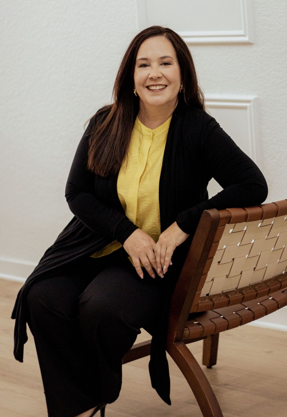 Professional woman wearing yellow blouse and black cardigan sits on wooden chair against white wall.