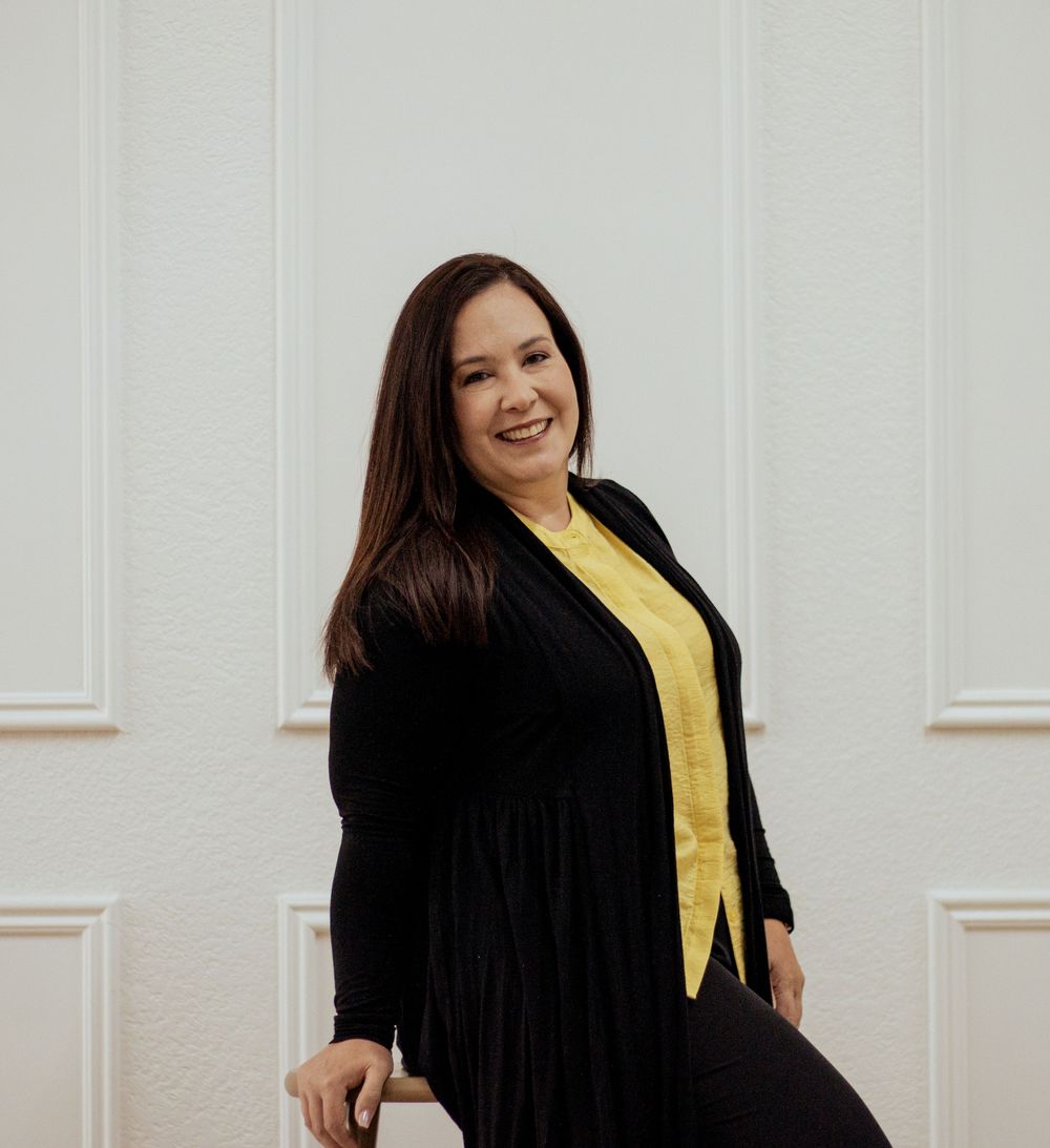 Woman in yellow top and black cardigan poses confidently against white wall with cheerful expression.