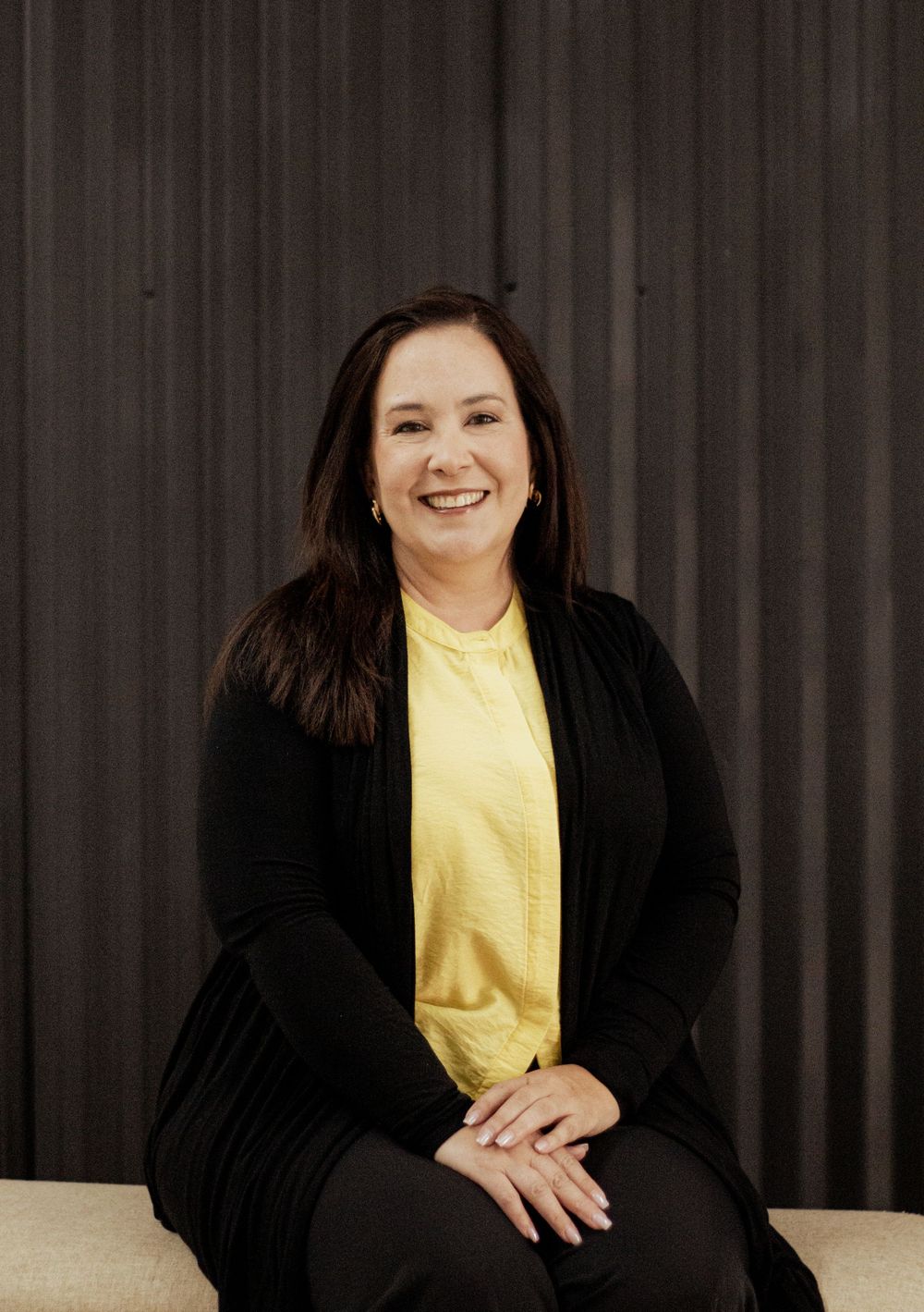 Professional portrait against dark backdrop showing person in yellow blouse and black cardigan seated with hands clasped.