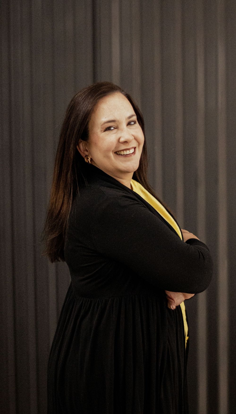 Professional portrait of someone in a black dress and yellow accent smiling warmly against a dark curtained background.