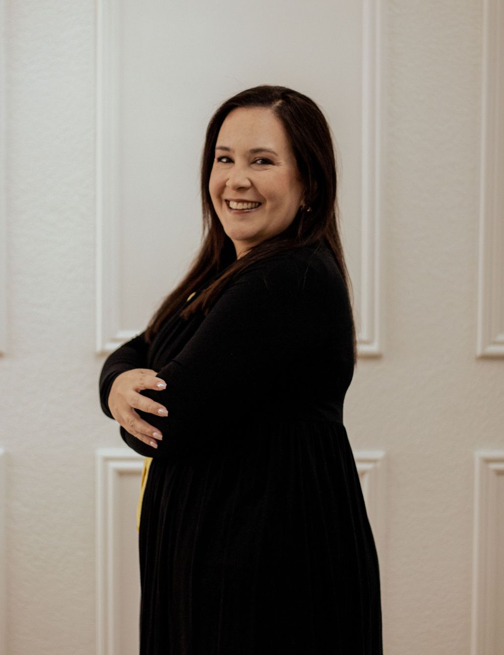 A woman in a black sweater poses against a white wall with architectural molding details while smiling warmly.