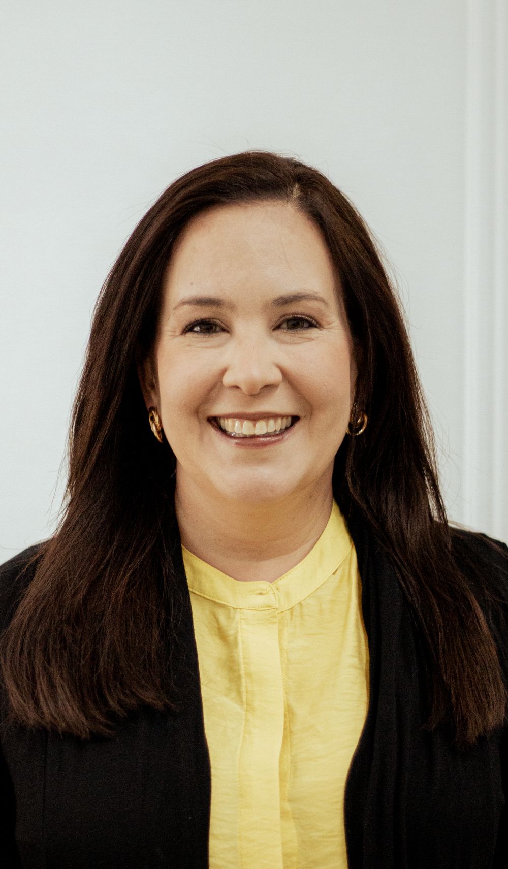 Professional headshot against white background wearing black blazer and yellow blouse, with warm smile.