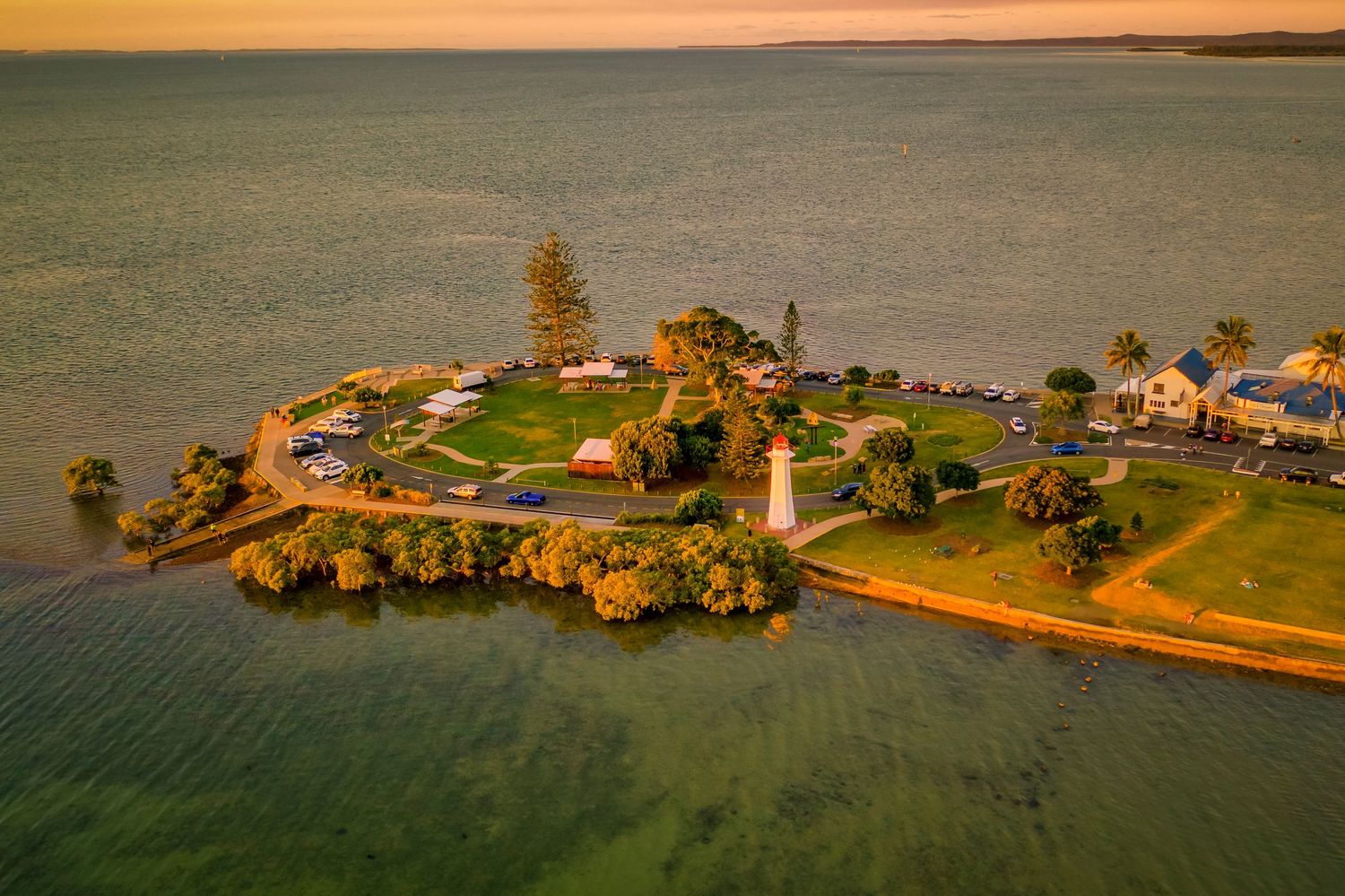 Aerial view of a coastal lighthouse and buildings on a small peninsula during golden hour.