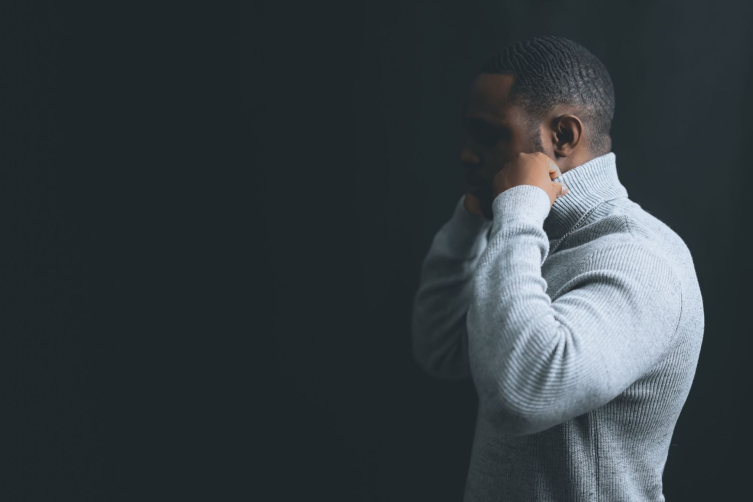 A person wearing a gray turtleneck sweater stands against a dark background with their hand covering their face.