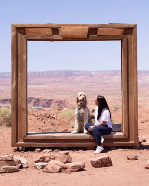 A wooden picture frame installation in the desert landscape overlooks scenic red rock cliffs and canyons with two figures inside.