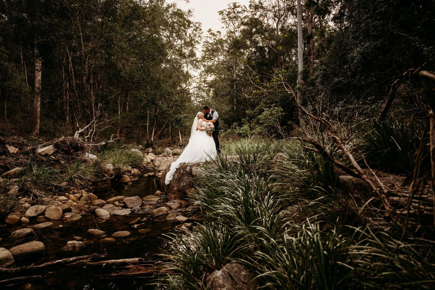 A couple in wedding attire embraces on rocky terrain surrounded by lush forest and native grasses at sunset.
