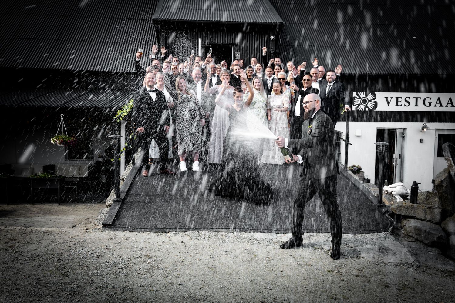 Wedding party celebrates outside Vestgaard venue with champagne spray in dramatic rain shower.