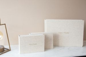Three elegant off-white linen photo albums of varying sizes displayed on a marble surface against a beige wall.