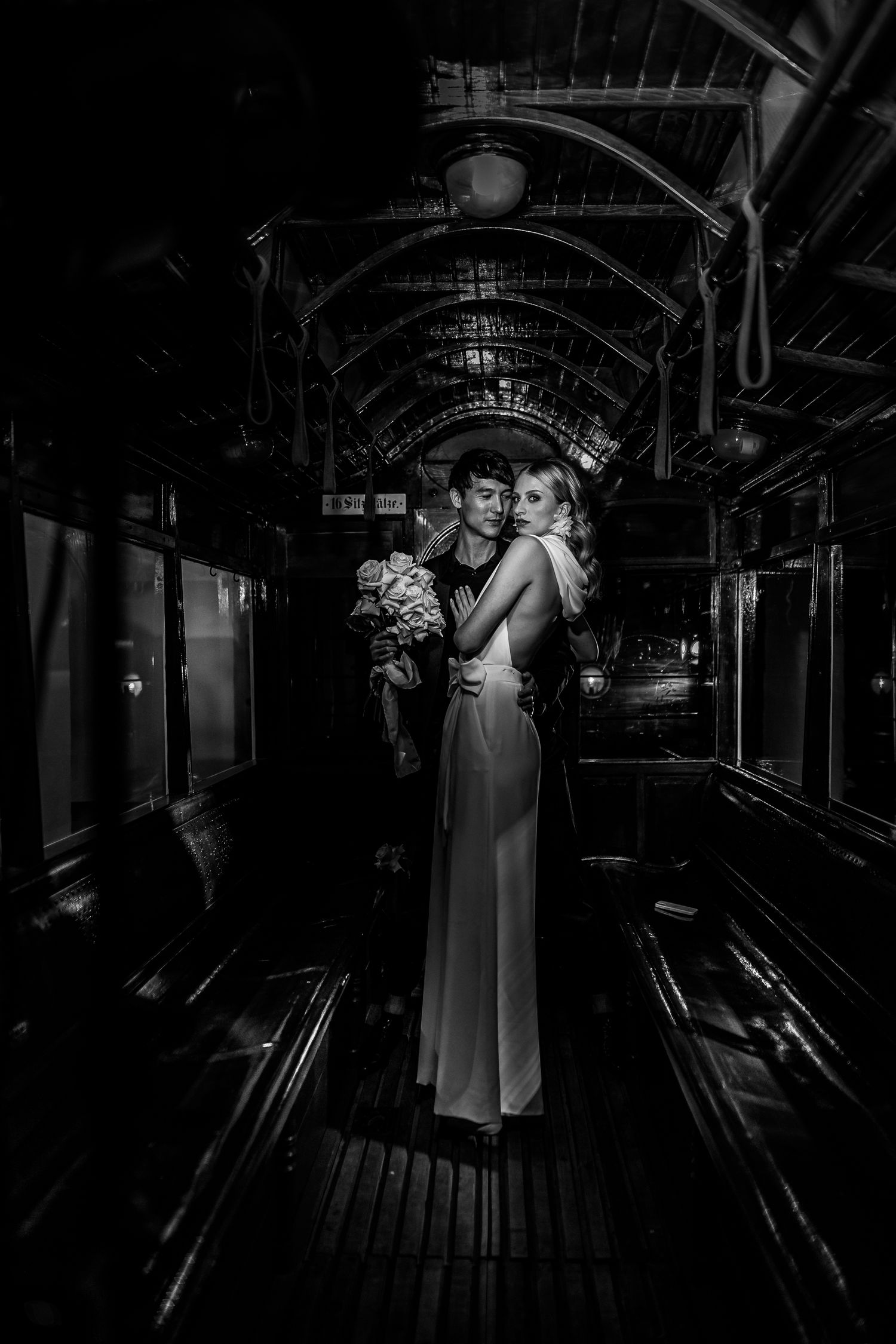 A grayscale image showing figures in white dresses walking through a dark, industrial hallway.