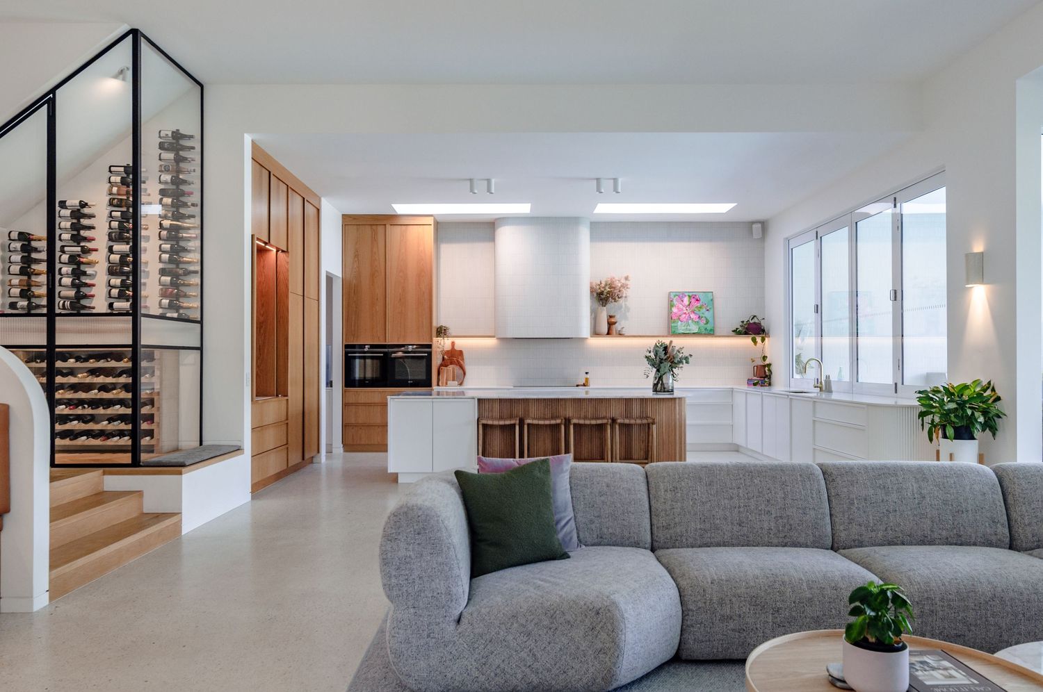 Modern living room features a grey sectional sofa, white walls, and natural light from large windows connecting open spaces.