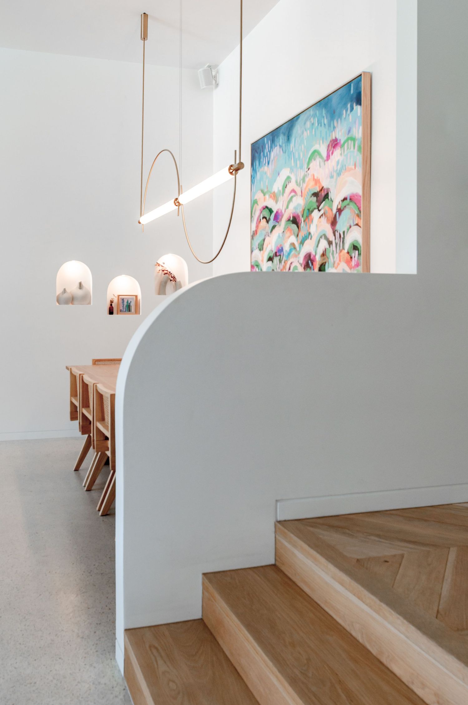 Modern interior space with curved white walls, wooden stairs, copper pendant lights and colorful abstract artwork on display.