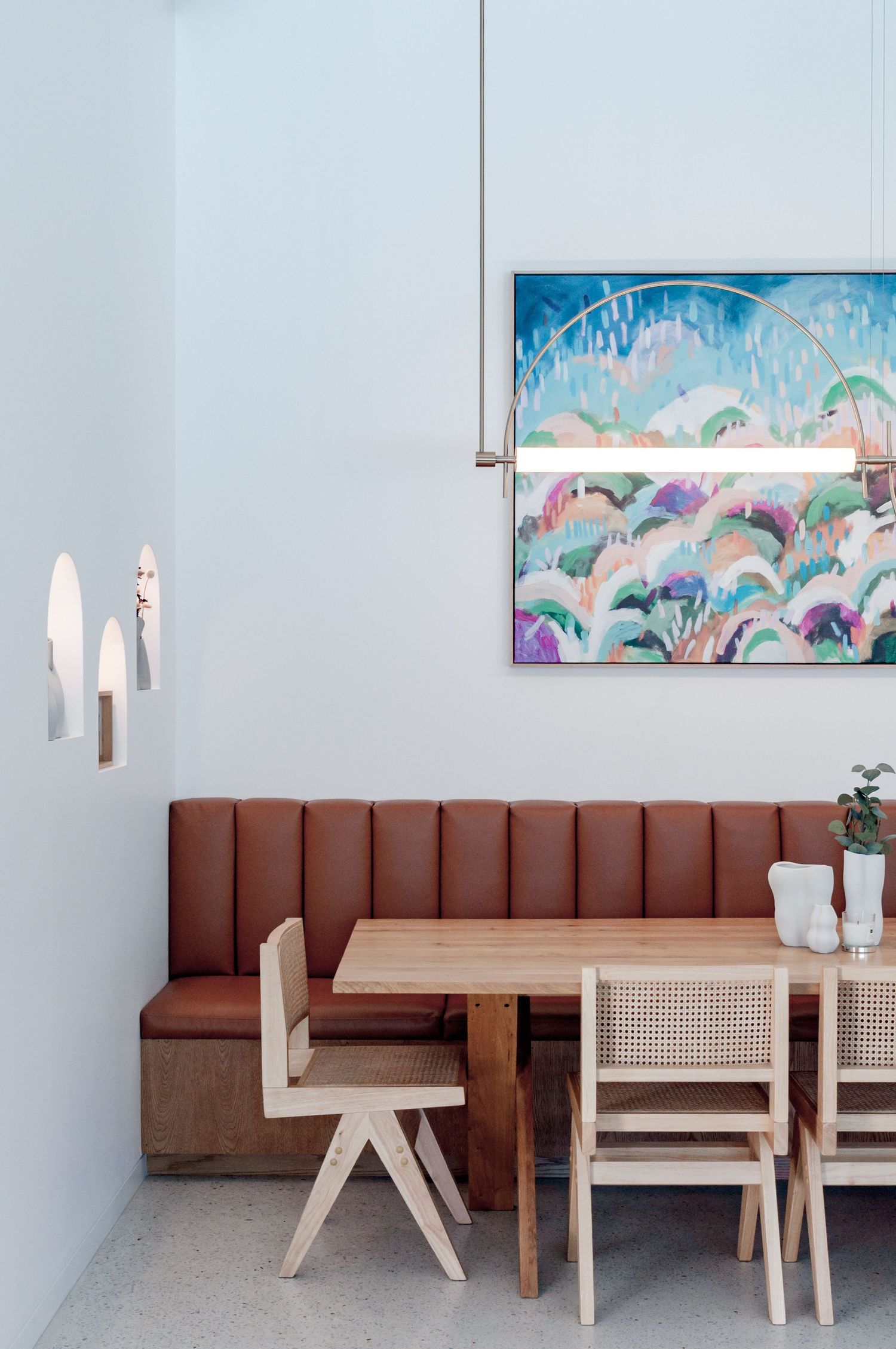 Modern restaurant seating with brown leather banquette, wooden chairs and colorful abstract artwork on white walls.
