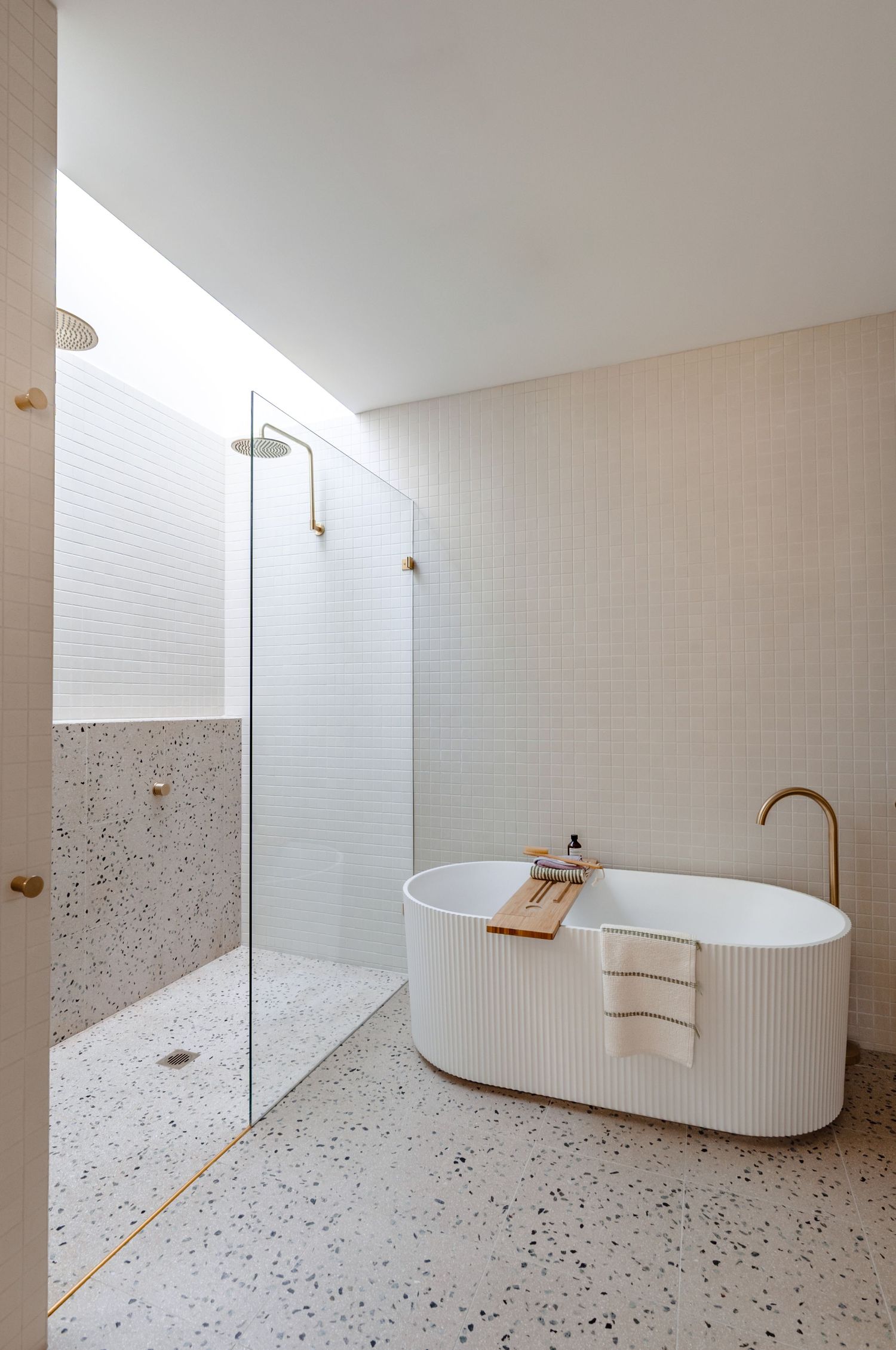 Modern minimalist bathroom with textured white bathtub, glass shower enclosure, terrazzo flooring and brass fixtures.