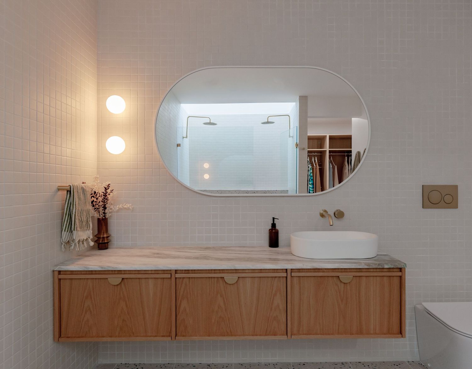 Modern bathroom design featuring floating wooden vanity, oval mirror, vessel sink and white tiled walls with warm lighting.