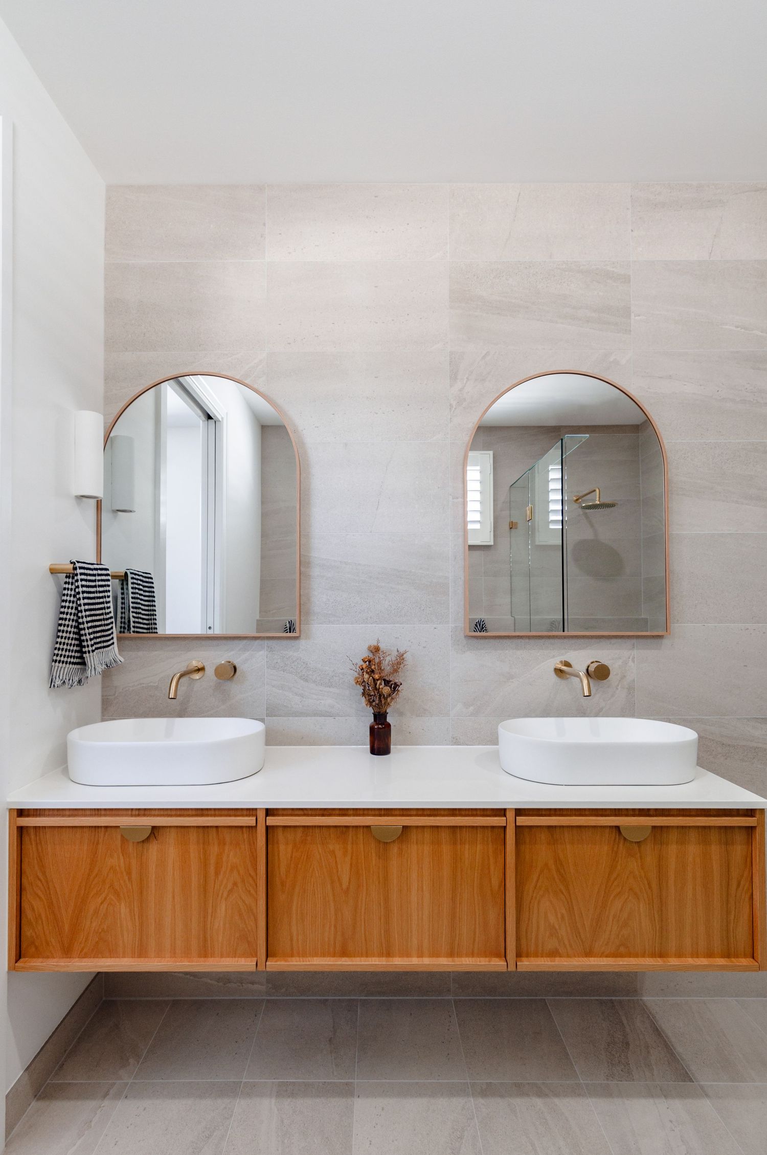 Modern bathroom featuring a floating wooden vanity with dual vessel sinks and arched mirrors against light stone walls.
