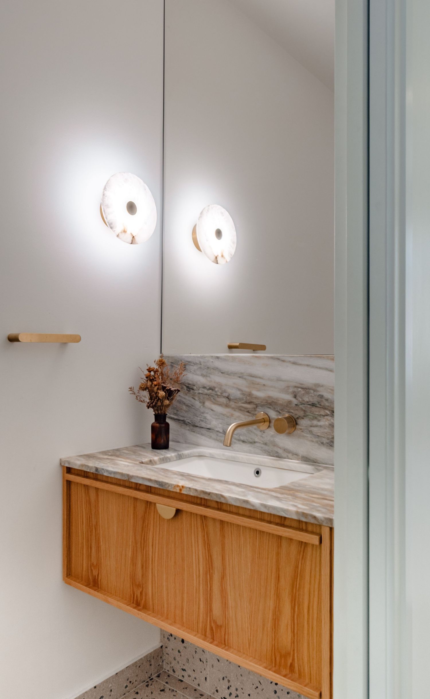 Modern bathroom vanity with floating wooden cabinet, marble countertop, gold fixtures and circular wall sconces.