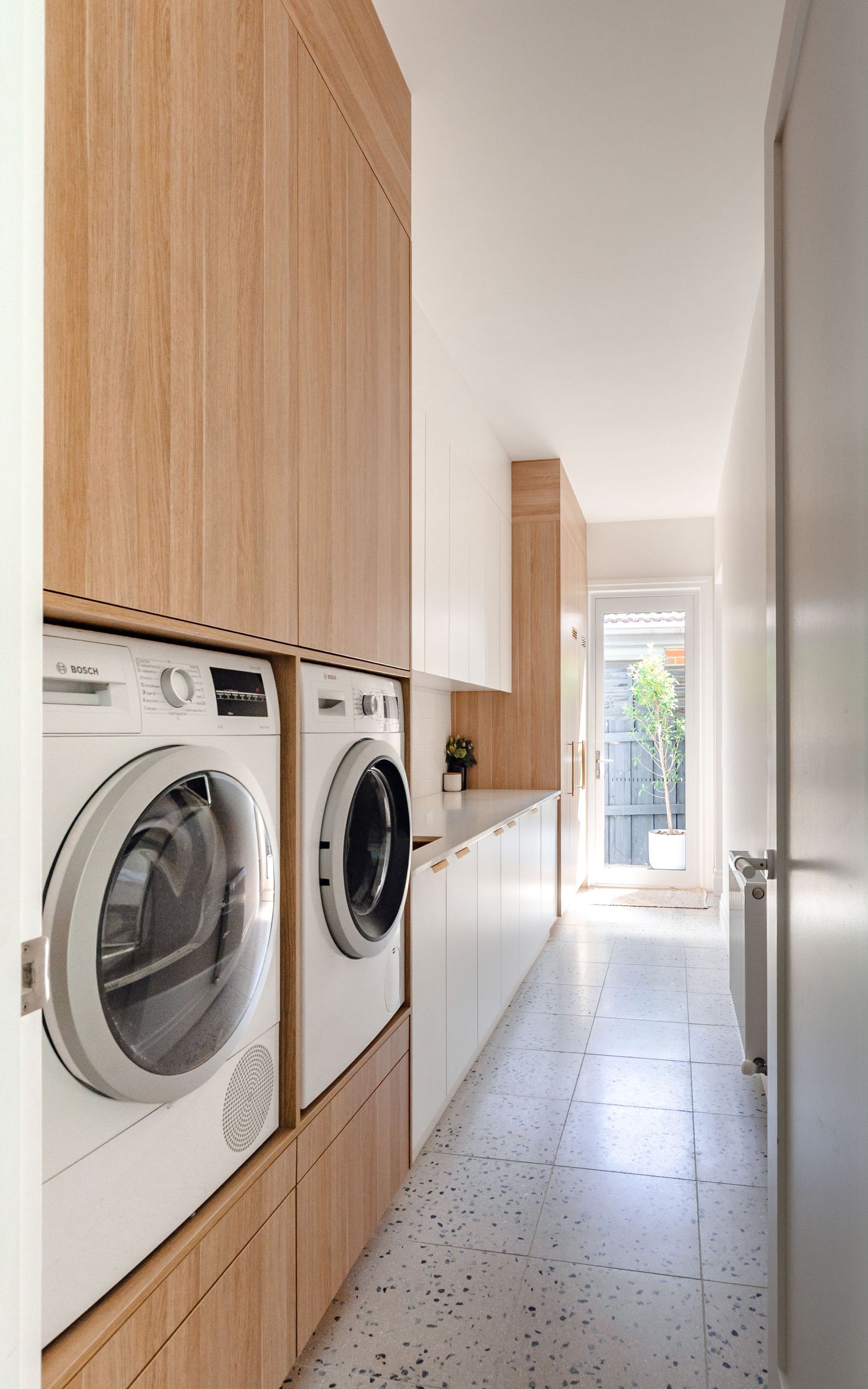 Modern laundry room with white appliances, wooden cabinets, and terrazzo flooring in a sleek minimalist corridor.