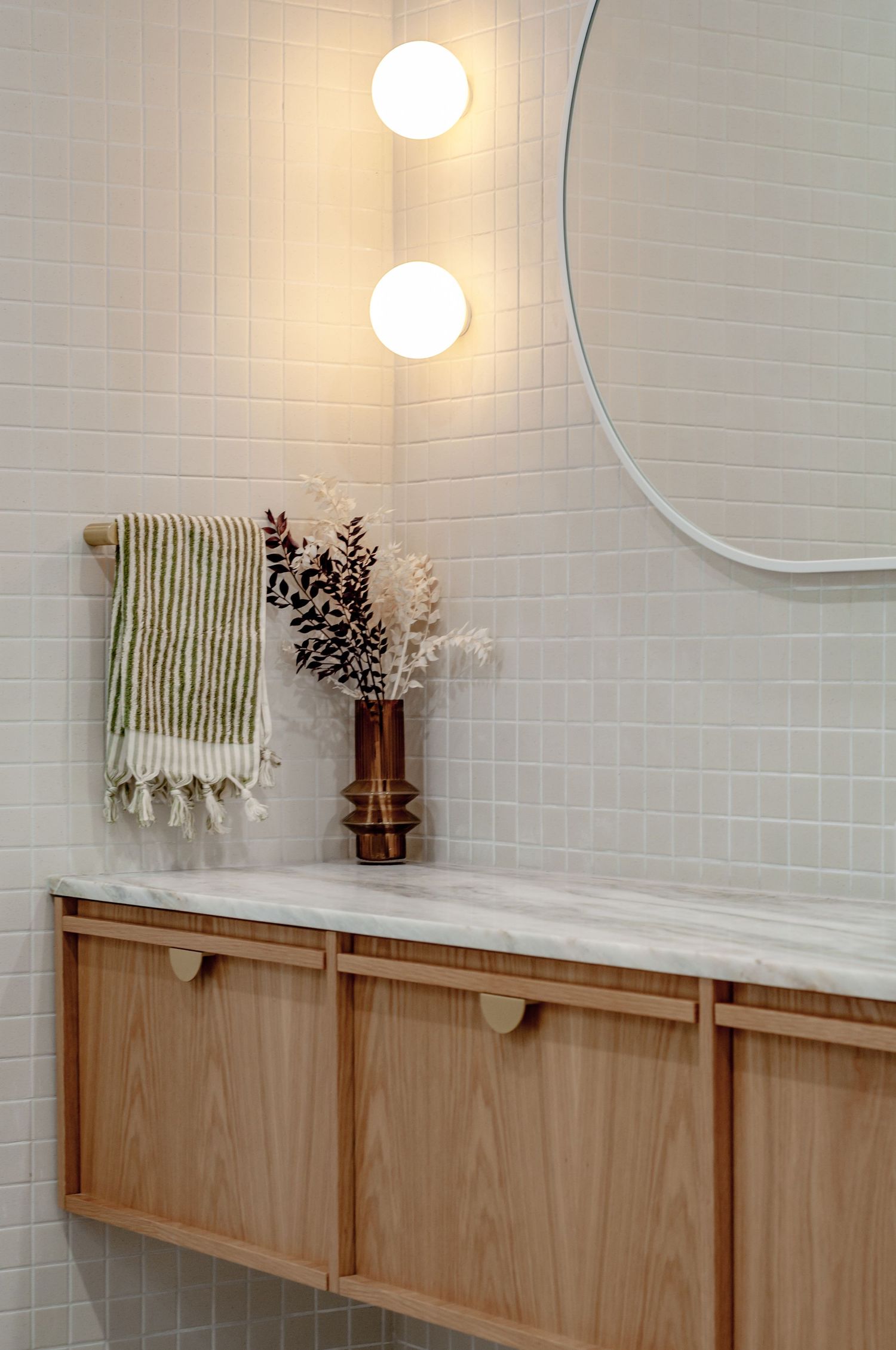 Minimalist bathroom vanity with light wood cabinets, round mirror, and globe wall sconces against white tile.