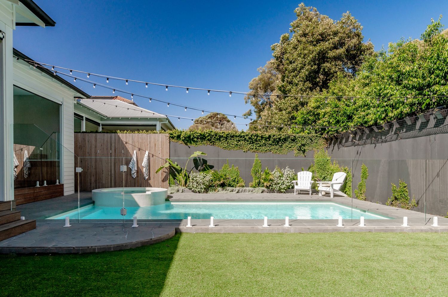 Modern backyard oasis featuring a rectangular swimming pool, artificial turf lawn, and string lights against blue sky.