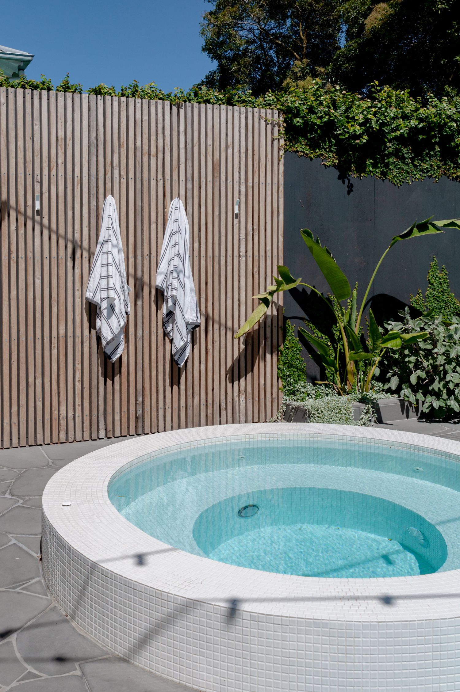 Modern outdoor hot tub with white mosaic tiles and wooden slatted wall featuring hung towels beside tropical plants.