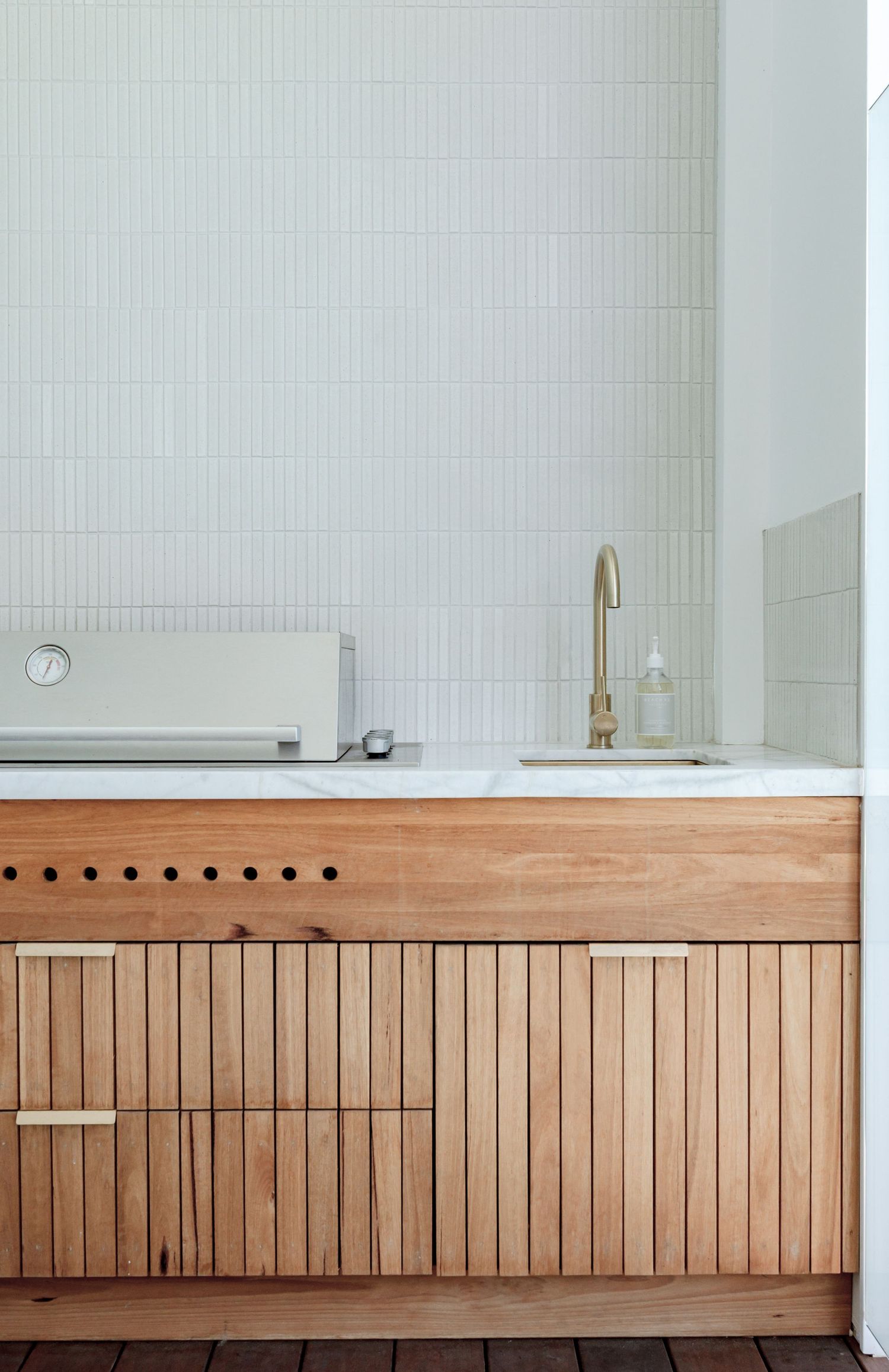 Minimalist wooden kitchen cabinetry with vertical slats and brass faucet against white tiled wall.