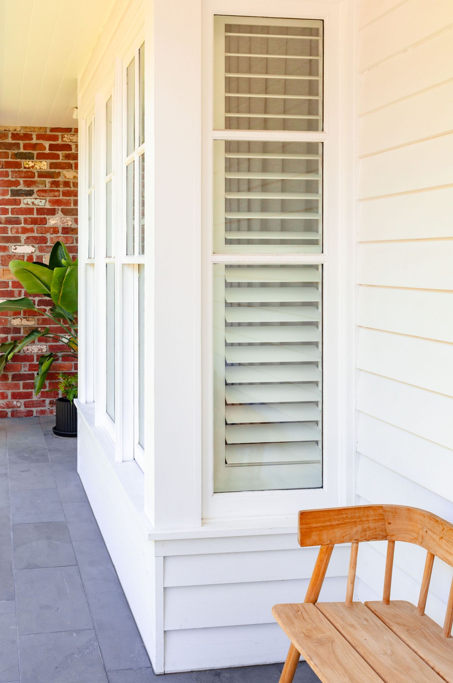 White louvered windows with cream siding and wooden chairs create a serene porch setting with exposed brick accents.