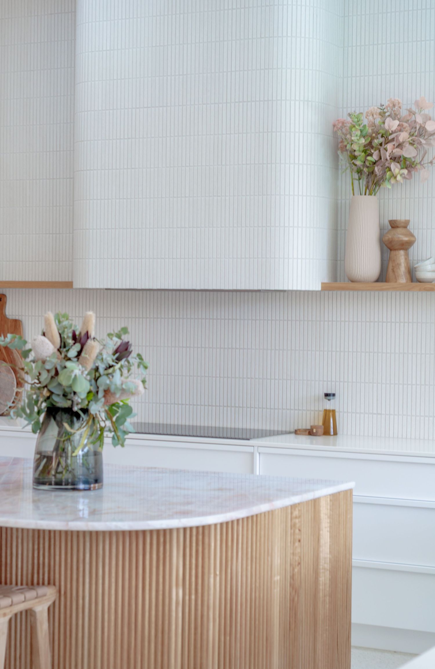 Modern kitchen interior with fluted wood island base, marble countertop, vertical tile backsplash and decorative floral arrangements.