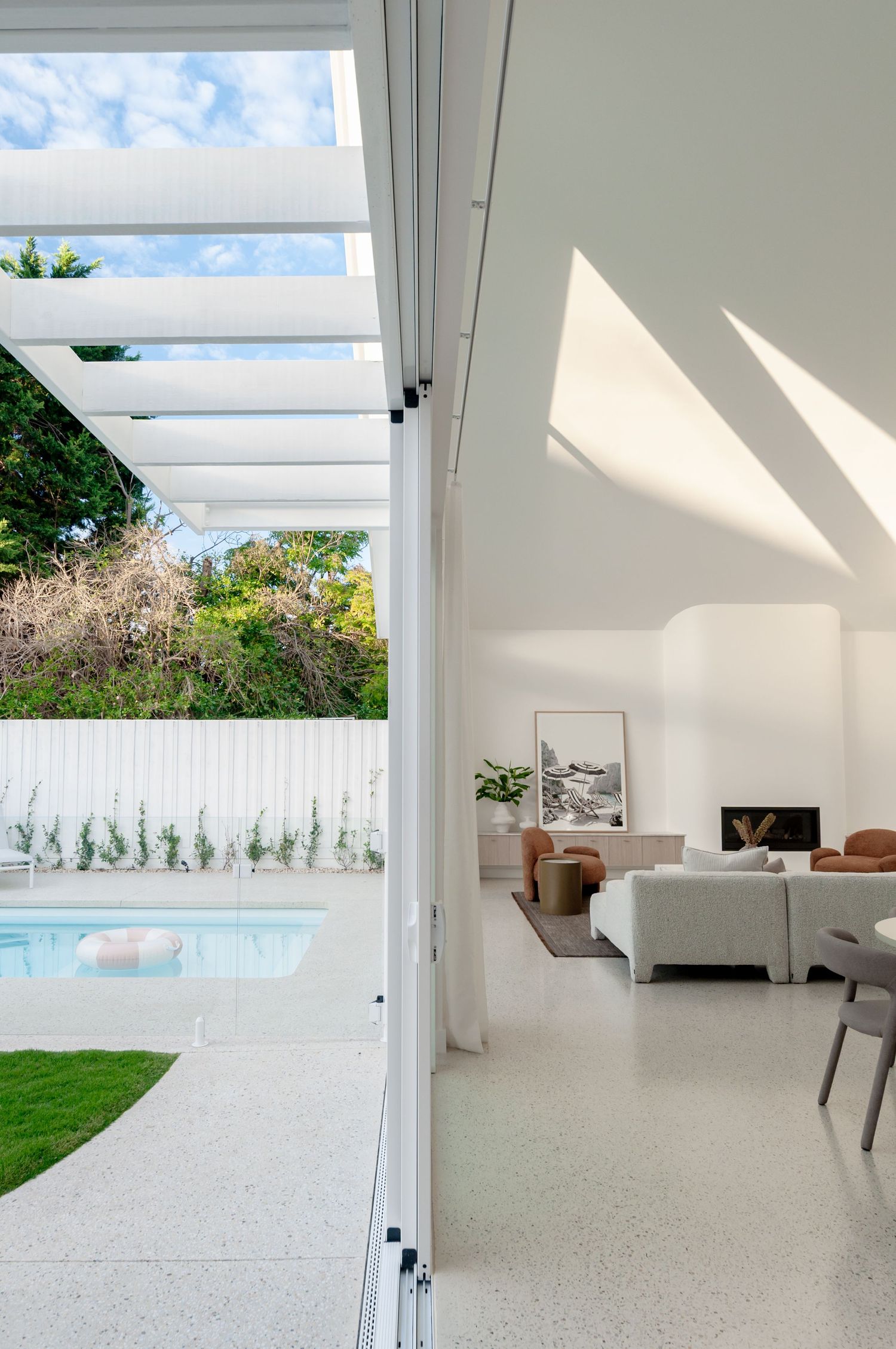 Minimalist white interior space with floor-to-ceiling windows casting dramatic shadows and views of outdoor landscaping.