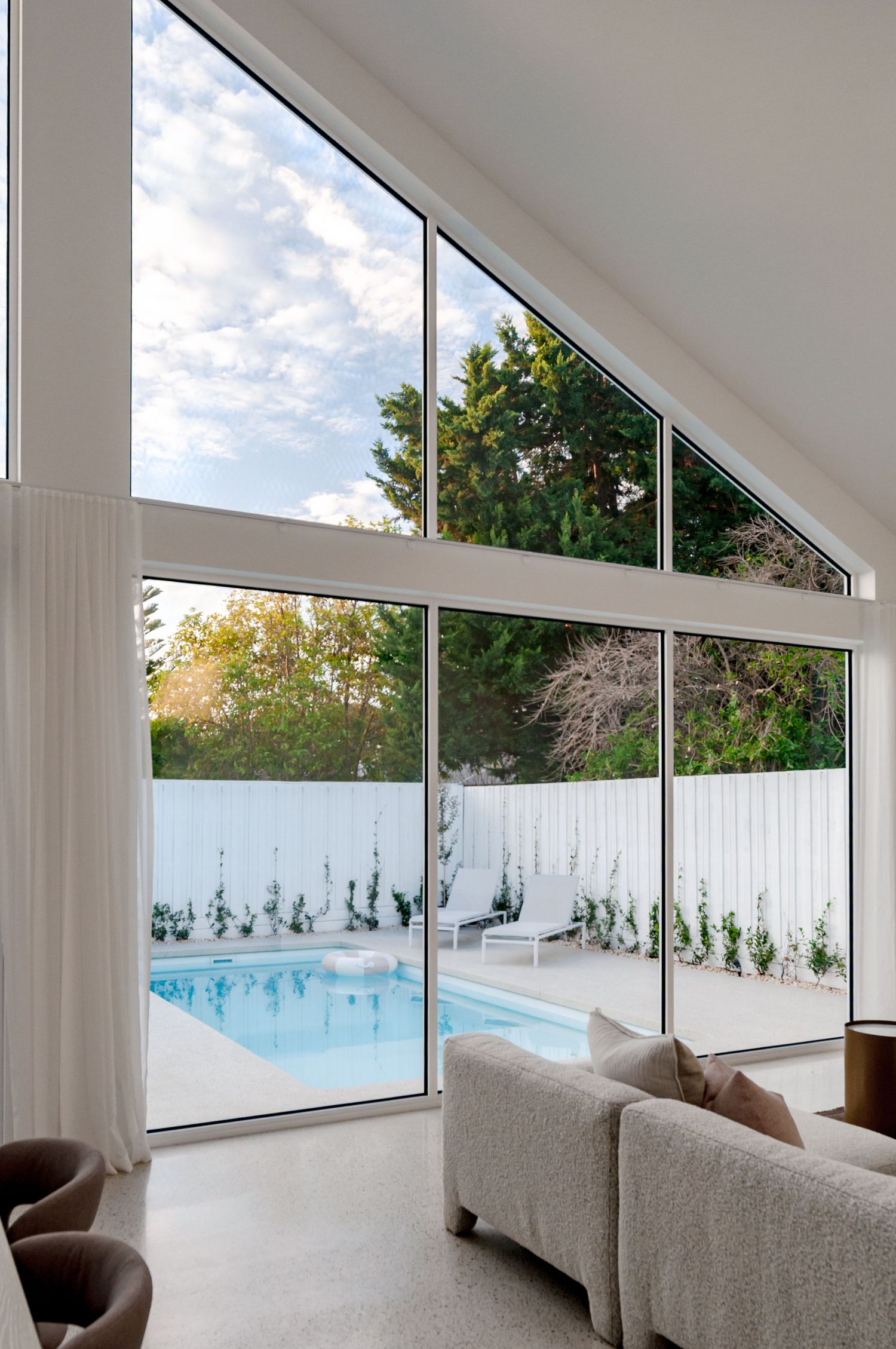 Contemporary living space with angled floor-to-ceiling windows overlooking a pool and white fence backyard.