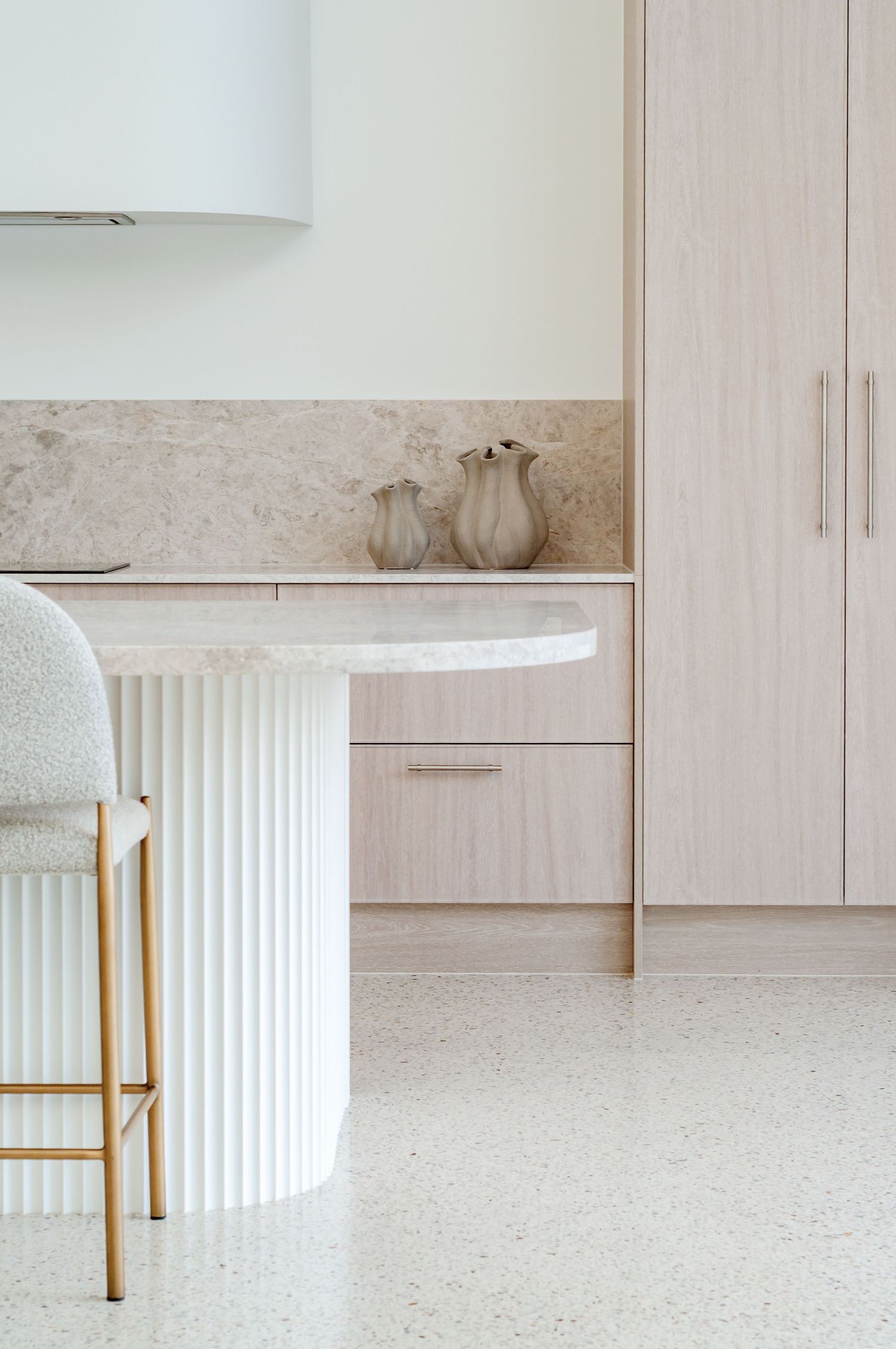 Close-up of minimalist kitchen detail showing fluted island base, white barstool and light wood cabinetry.