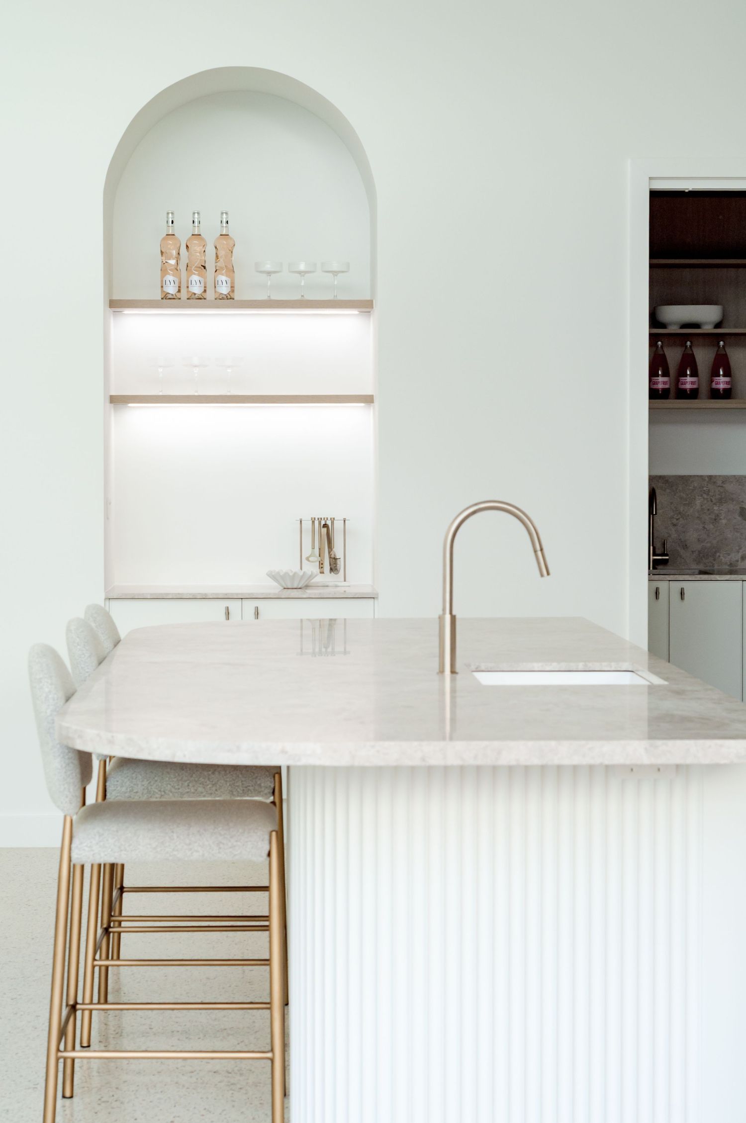Modern white kitchen with marble countertop, brass faucet, and arched alcove shelving displaying glass bottles.