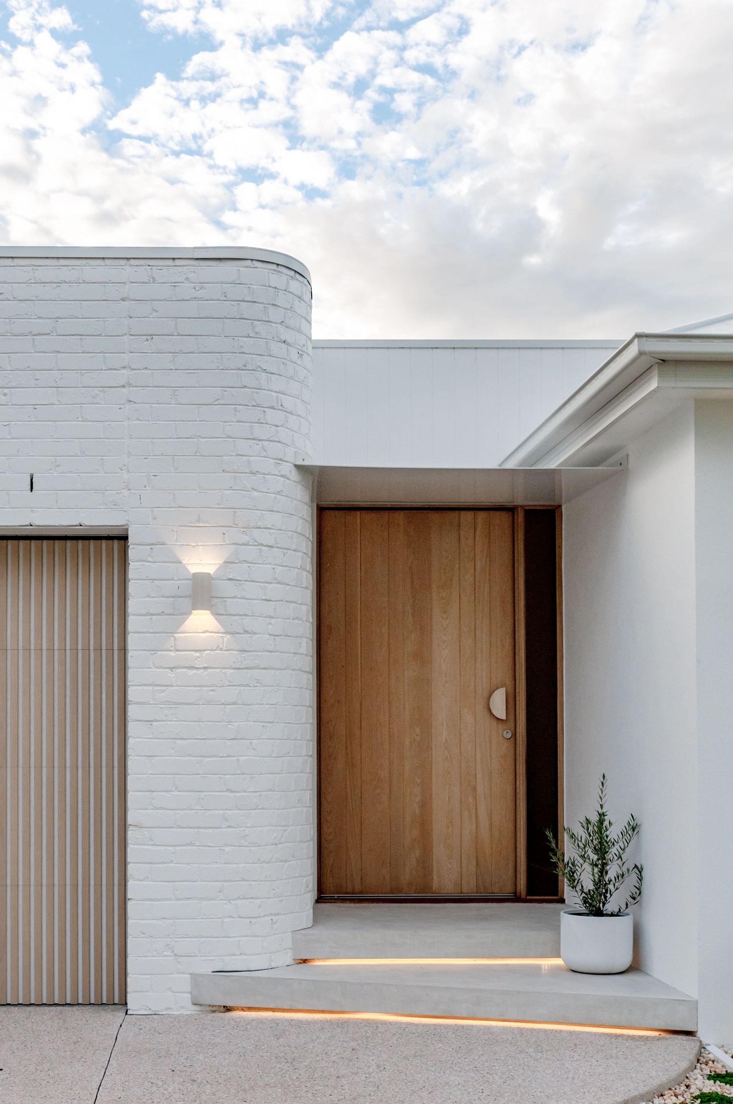 Modern white brick home entrance featuring wooden door, curved wall detail and wall-mounted accent lighting.