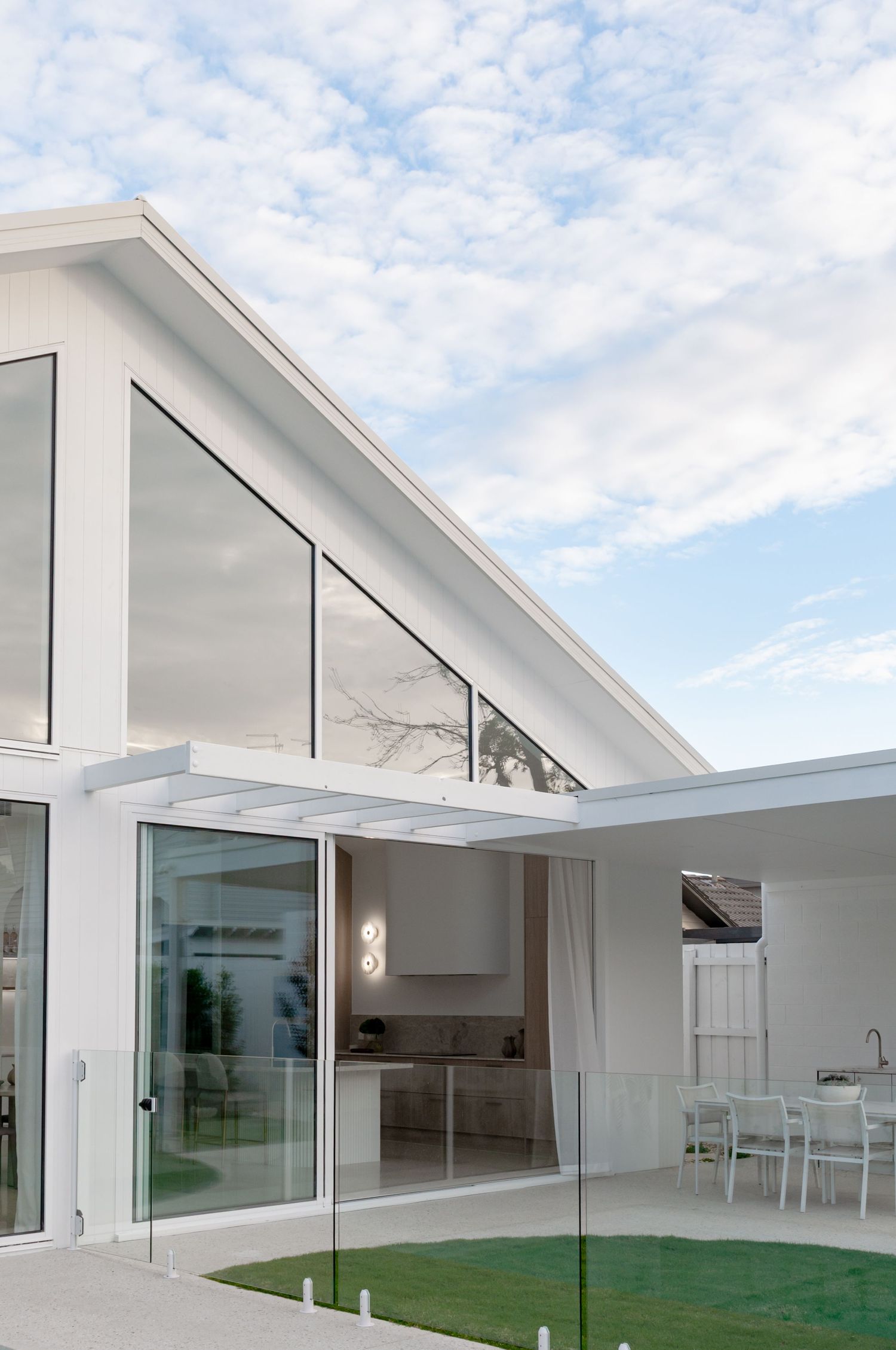 Modern white exterior architecture features angular rooflines and glass walls overlooking a green lawn under cloudy skies.