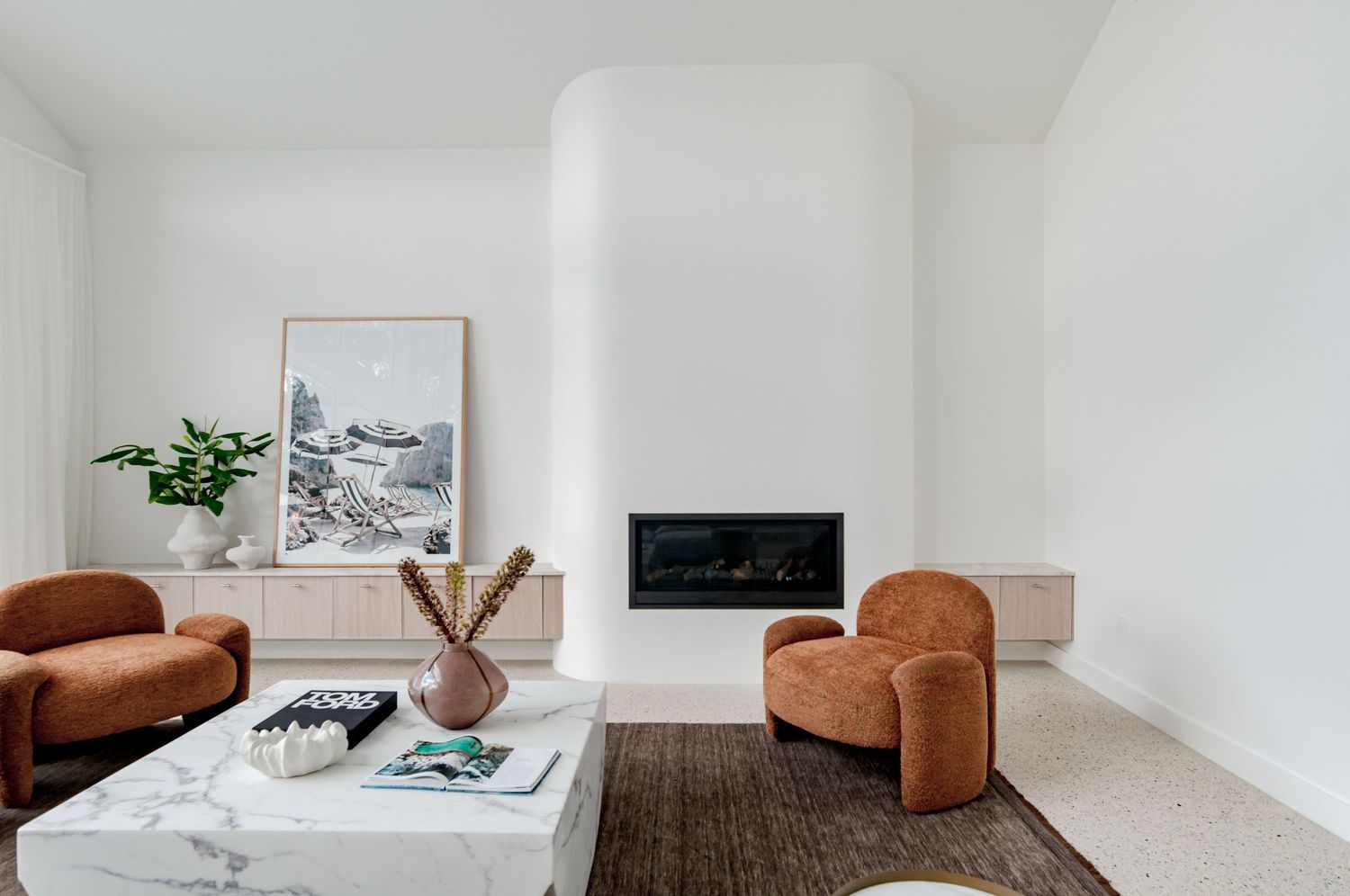 Minimalist living room with brown curved armchairs, white walls, and a built-in fireplace with modern artwork above.