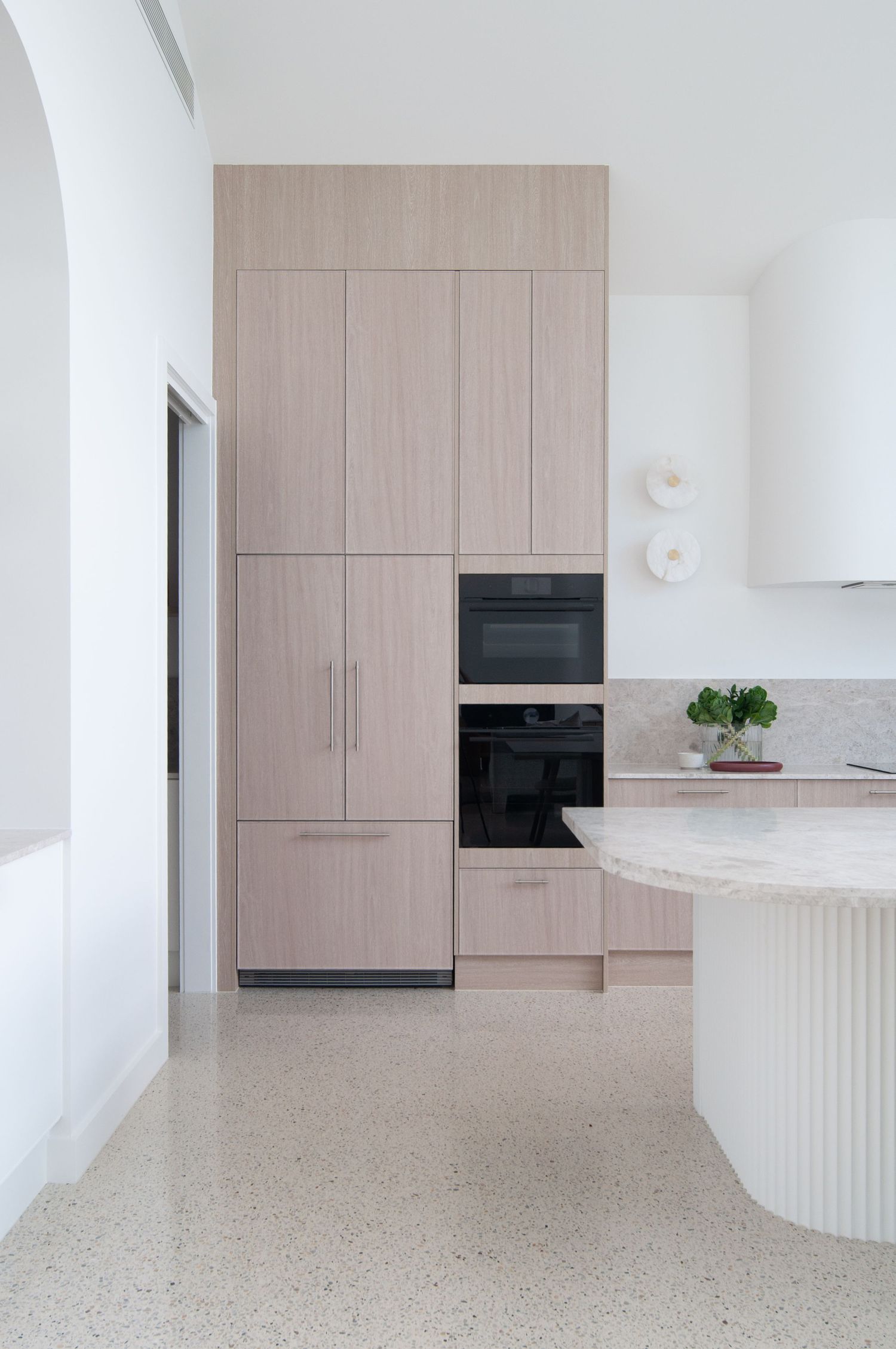 Modern kitchen featuring light wood cabinetry, built-in appliances, and a curved white island with terrazzo flooring.