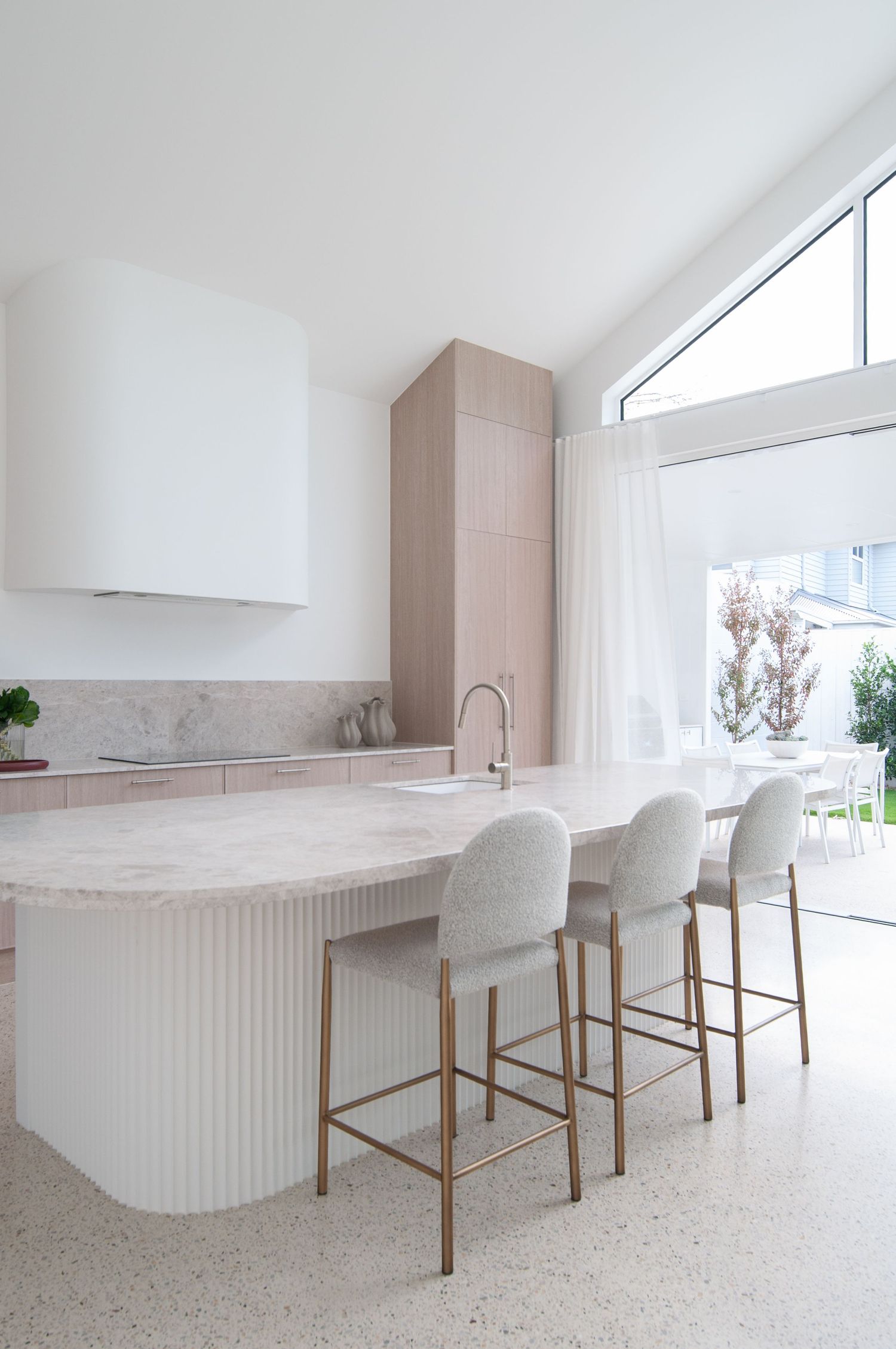 Modern white kitchen featuring a curved marble island with textured paneling and three upholstered barstools.