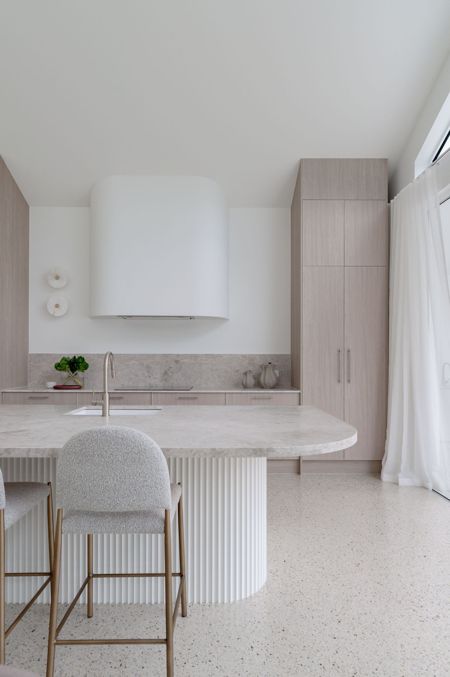 Modern minimalist kitchen featuring a curved marble island with fluted base, textured bar stool, and white range hood.